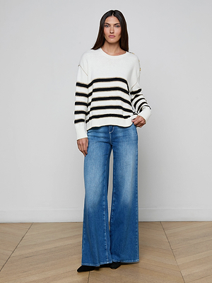 A woman with long brown hair and a neutral expression stands indoors in a minimal setting, wearing the Bishop Striped Sweater with black horizontal stripes and gold buttons, paired with wide-leg blue jeans and black shoes.