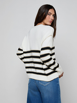 A woman with long dark hair, wearing the Bishop Striped Sweater with black stripes and blue jeans, stands with her back turned and looks over her shoulder against a plain light background.