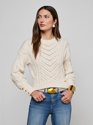 A woman models the L'AGENCE Storm Cable-Knit Sweater, featuring a cream chevron pattern and gold buttons, paired with blue jeans and gold accessories. She stands against a light gray background, looking to her left.