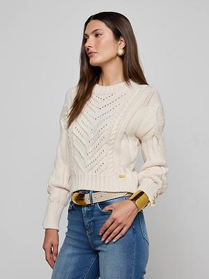 A woman with long brown hair poses against a light gray background, wearing the L'AGENCE Storm Cable-Knit Sweater in cream with blue jeans, gold earrings, a gold bracelet, and a beige belt.