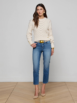 A woman with long brown hair and a neutral expression stands against a plain light background, wearing the L’AGENCE Storm Cable-Knit Sweater in cream with gold buttons, paired with blue jeans, beige heels, gold earrings, and a gold belt.