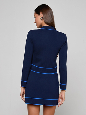 A woman with straight brown hair faces away, wearing the Tala Cardigan in navy blue with light blue trim. She poses against a plain light gray background.