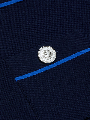 A close-up of the Cecilia Knit Mini Skirt features a navy blue fabric pocket with a silver eagle button, decorative text, and two thin blue stripes above and below, highlighting refined simplicity and subtle contrast piping.
