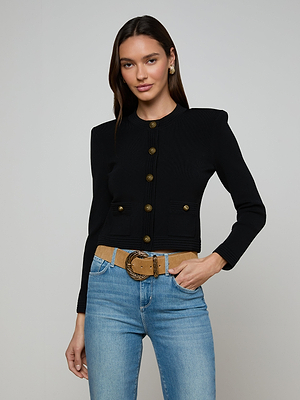 A woman with long brown hair wears the Patsy Cropped Cardigan, a fitted black top with signature buttons, paired with light blue jeans and a tan belt. She stands against a plain light gray background, smiling softly.