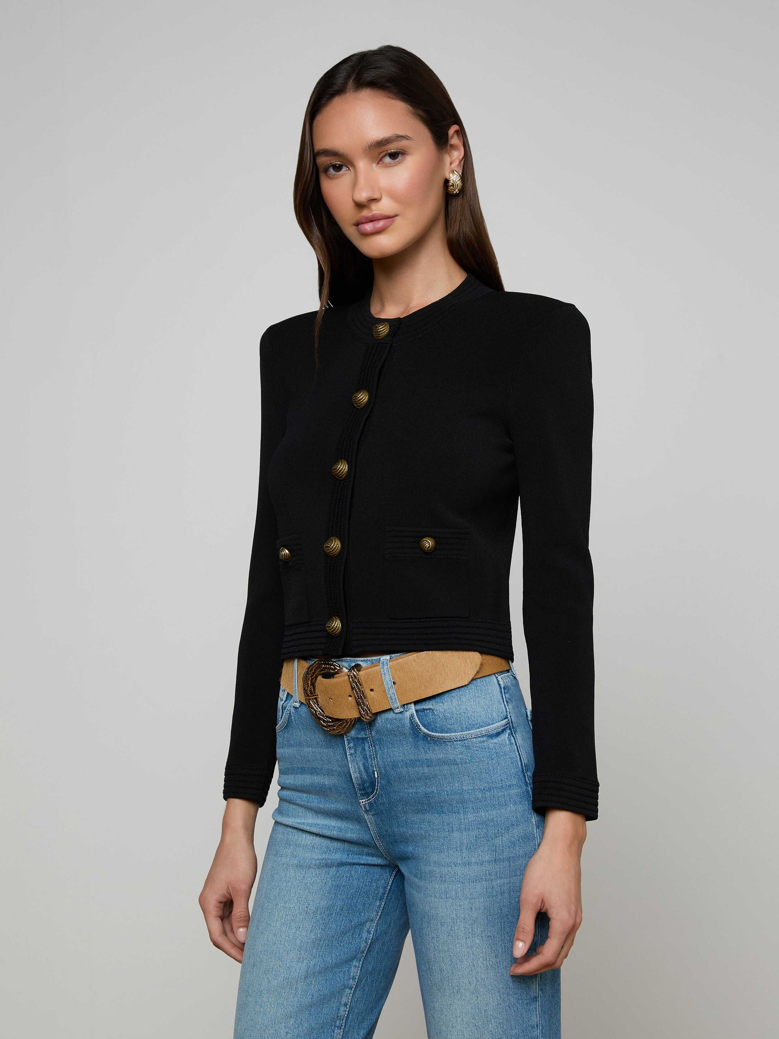 A woman wearing the Patsy Cropped Cardigan in black with signature buttons, paired with light blue jeans and a tan belt, stands against a plain light background.