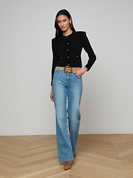 A woman poses against a plain white background wearing the Patsy Cropped Cardigan in black with signature gold buttons, paired with light blue high-waisted jeans, a wide belt, and tan heels.