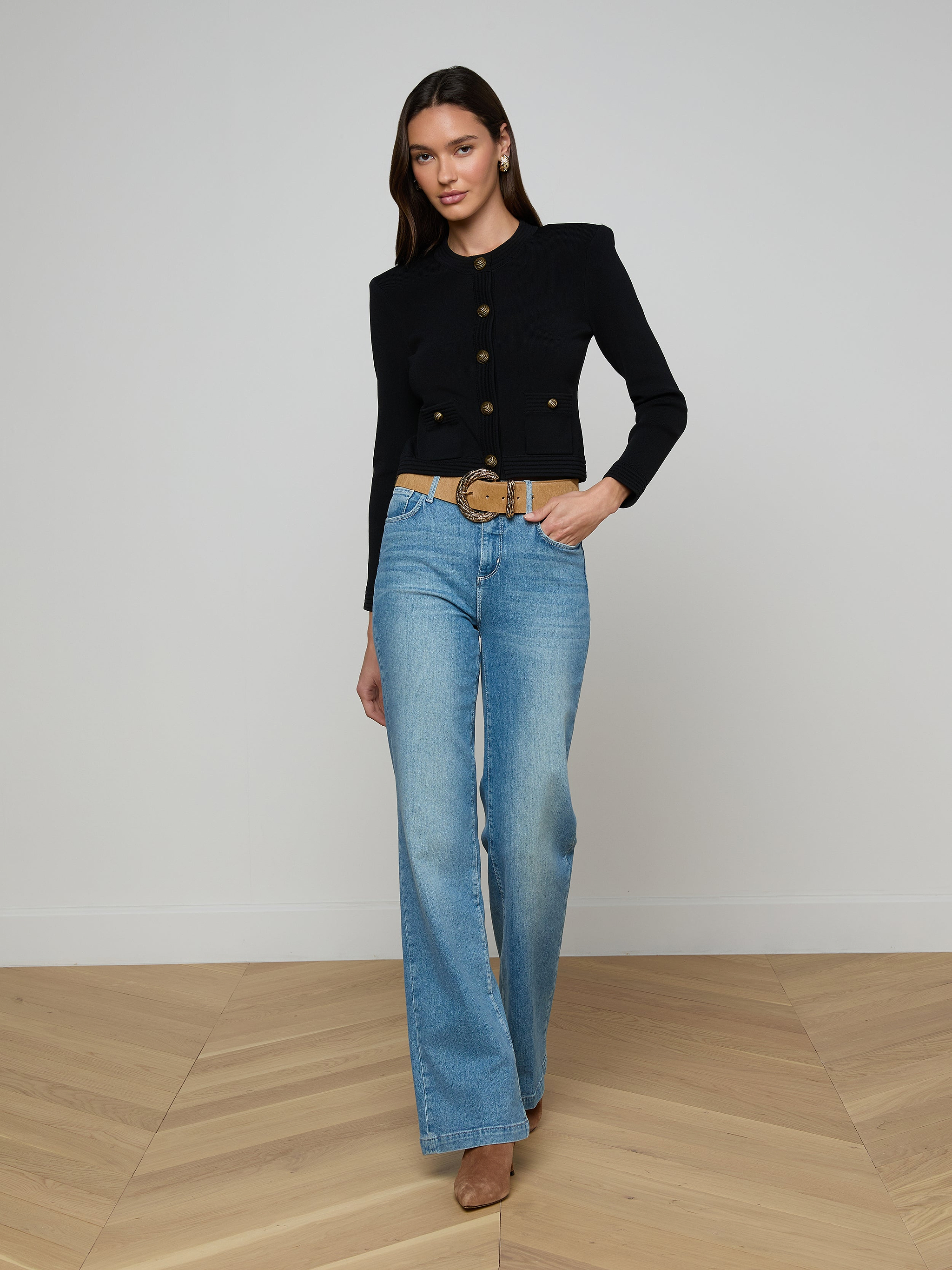 A woman poses against a plain white background wearing the Patsy Cropped Cardigan in black with signature gold buttons, paired with light blue high-waisted jeans, a wide belt, and tan heels.