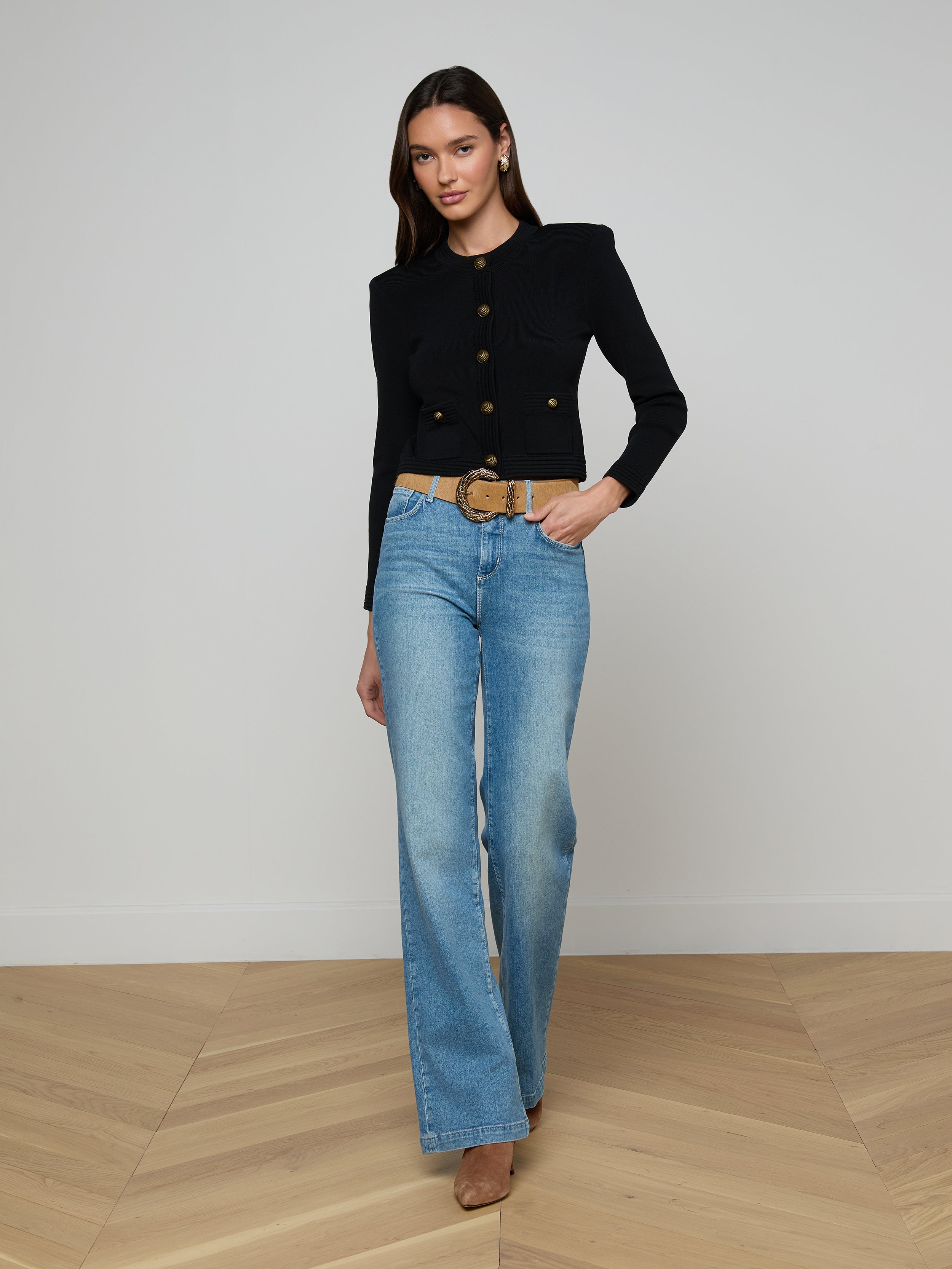 A woman poses against a plain white background wearing the Patsy Cropped Cardigan in black with signature gold buttons, paired with light blue high-waisted jeans, a wide belt, and tan heels.