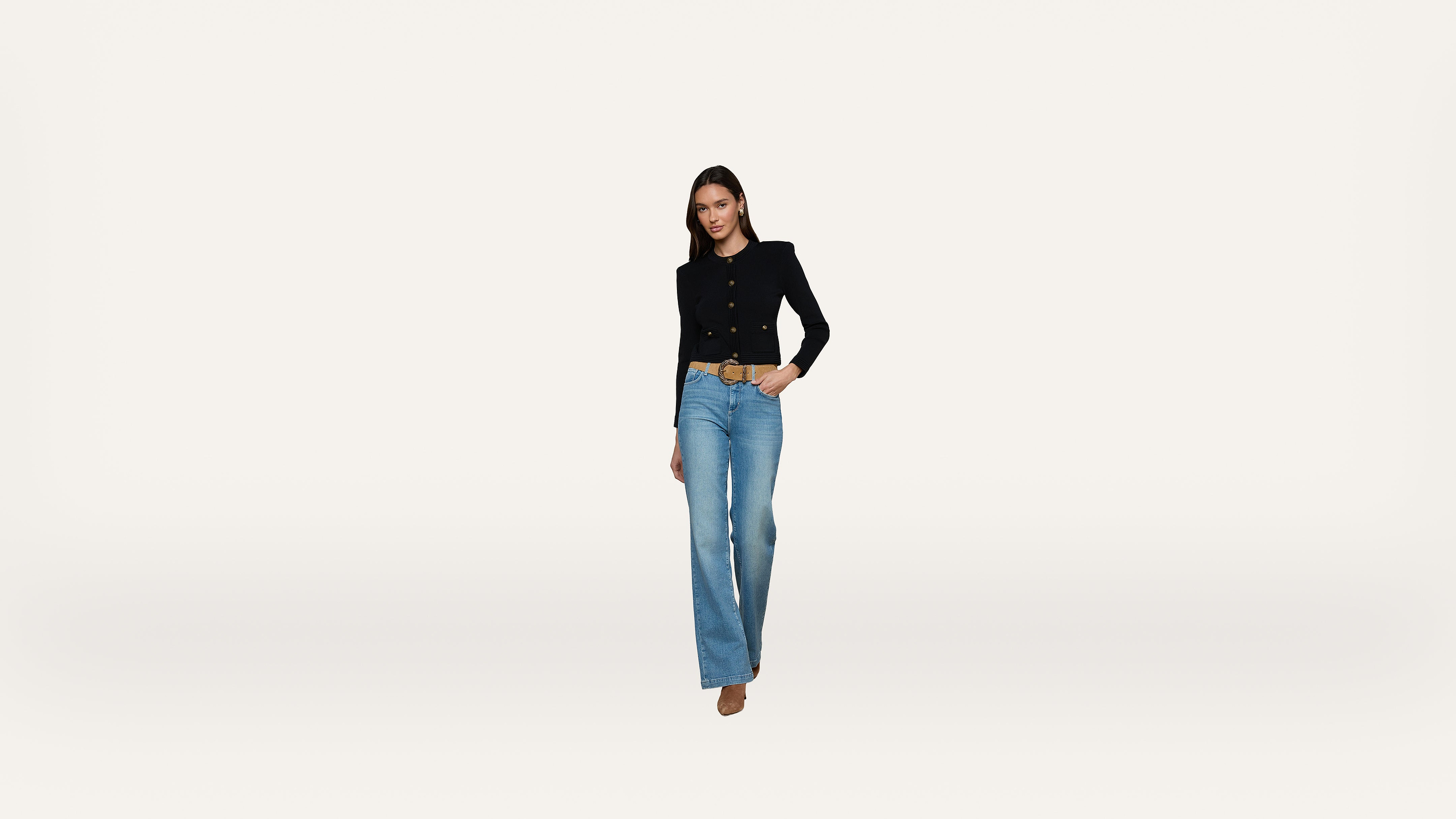 A woman stands against a plain white background wearing a black blazer, blue jeans, and brown shoes, with one hand in her pocket and a slight smile.