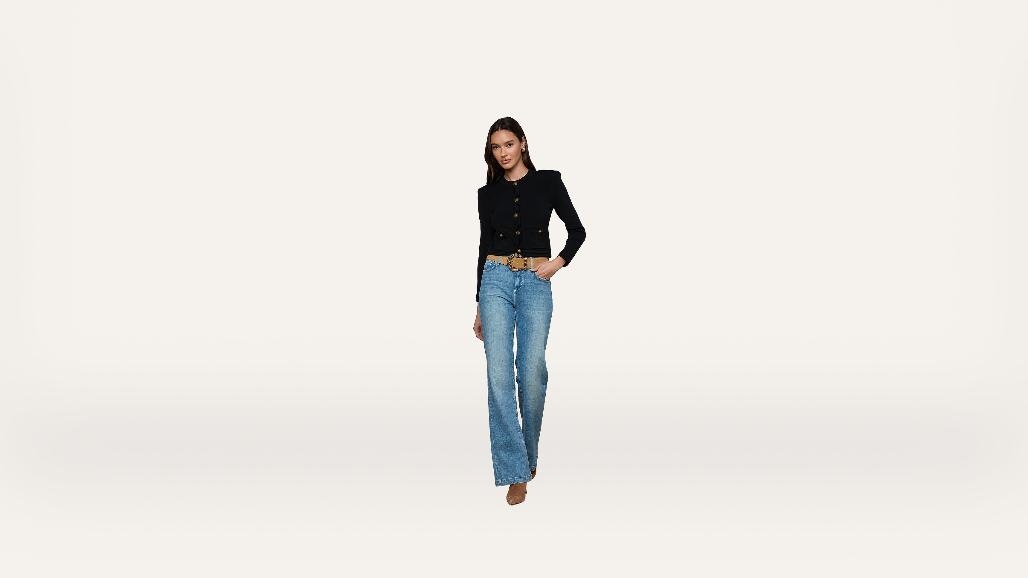 A woman stands against a plain white background wearing a black blazer, blue jeans, and brown shoes, with one hand in her pocket and a slight smile.
