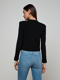 A woman stands facing away, wearing the Patsy Cropped Cardigan—a black power knit long-sleeve top with signature buttons—paired with light blue jeans. Her straight brown hair is down, and both hands rest by her sides against a plain, light background.