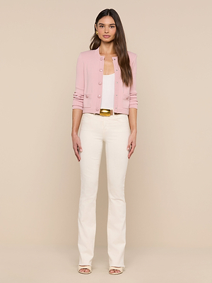A woman faces forward against a plain beige background, wearing the Patsy Cropped Cardigan in light pink over a white top, paired with cream pants and open-toe heels.