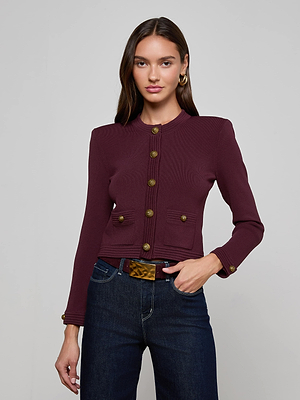 A woman with long brown hair wears the L'AGENCE Patsy Cropped Cardigan, featuring gold signature snap buttons, paired with dark blue jeans and a brown belt, standing against a plain light gray background.