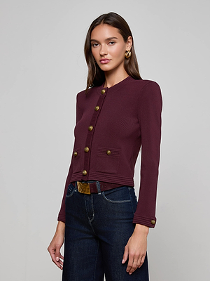 A woman with long brown hair models the L'AGENCE Patsy Cropped Cardigan, featuring signature snap buttons and ribbed trims, paired with dark blue jeans against a plain light gray background as she looks confidently at the camera.