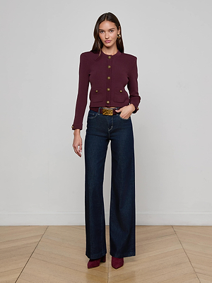 A woman stands indoors against a white wall and wooden floor, wearing the L'AGENCE Patsy Cropped Cardigan in burgundy, paired with high-waisted dark blue wide-leg jeans, a brown belt with gold buckle, and burgundy heels.