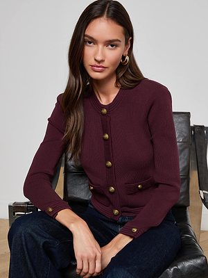 A woman with long brown hair sits on a black chair, wearing the L’AGENCE Patsy Cropped Cardigan with signature snap buttons, dark jeans, and gold hoop earrings, looking directly at the camera with a neutral expression.