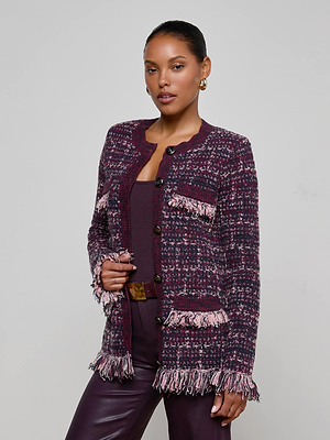 A woman stands against a plain background, confidently wearing the L'AGENCE Kastoria Fringe Cardigan—a textured knit in burgundy and pink—over a matching top and burgundy leather pants, accessorized with hoop earrings and holding her jacket lapel.