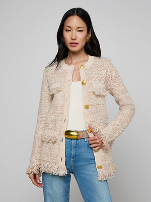 A woman with long dark hair and gold earrings wears the Kastoria Fringe Cardigan—cream boucle tweed with large gold buttons—in a longline silhouette, styled over a white top and blue jeans, standing against a plain light gray background.