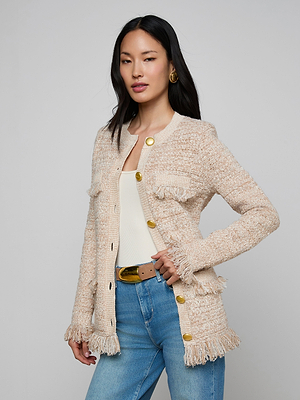 A woman stands confidently against a gray background wearing the Kastoria Fringe Cardigan, a textured knit beige piece with gold buttons, styled with a white top, blue jeans, tan belt, gold hoop earrings, and long dark hair.