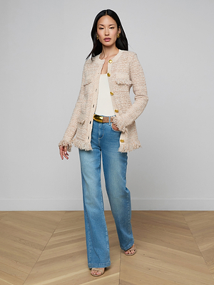 A woman stands indoors on a wooden floor, wearing the Kastoria Fringe Cardigan with gold buttons, a white top, blue high-waisted jeans, and beige heeled sandals. She has long dark hair and a neutral expression.