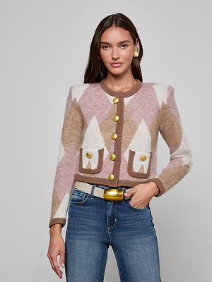 A woman with long brown hair and a neutral expression stands against a plain background wearing the Siobhan Cardigan—a statement argyle knit with gold buttons—styled with blue jeans, a beige belt, and gold earrings.
