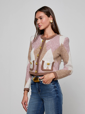 Wearing the Siobhan Cardigan in pink, brown, and white argyle with gold buttons, paired with blue jeans and gold earrings, she stands against a plain light background. Her statement knit layer and long brown hair add a chic touch to her look.