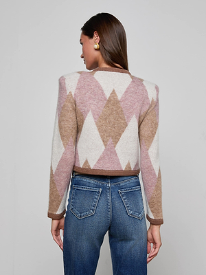 A woman with straight brown hair wears the Siobhan Cardigan, featuring an argyle knit in pink, brown, and white tones, layered over blue jeans. She stands with her back to the camera against a plain background.