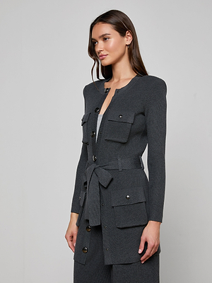 A woman with long brown hair wears the Lore Cardigan—a tailored dark gray knit sweater jacket with a tie belt, large flap pockets, and metallic buttons—standing against a plain light background.