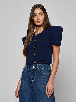 A woman with long brown hair wears the Britt Statement Shoulder Cardigan, a navy short-sleeve top with bold shoulder pads and large gold buttons, paired with high-waisted blue jeans against a plain light gray background.