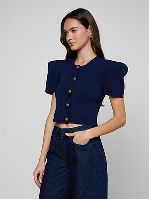 A woman stands against a plain background wearing the Britt Statement Shoulder Cardigan, a navy short-sleeve top with bold shoulder pads and large buttons, paired with high-waisted blue jeans. She poses looking slightly left, hands on her waist.