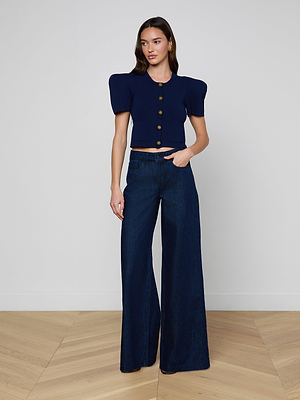 A woman with long brown hair stands indoors on a wood floor, wearing a navy blue short-sleeve top with large buttons and puffed shoulders, paired with the Pacey Wide-Leg Jean in a dark wash. She has a neutral expression.