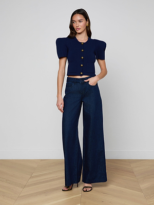 A woman with long hair and black heels poses confidently indoors in the Pacey Wide-Leg Jean and a navy blue, short-sleeved, buttoned crop top with structured shoulders against a plain wall.