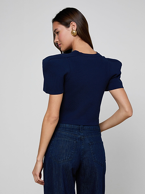 A woman with long brown hair wears the Britt Statement Shoulder Cardigan and high-waisted blue jeans, standing and facing away while looking over her left shoulder against a plain background.