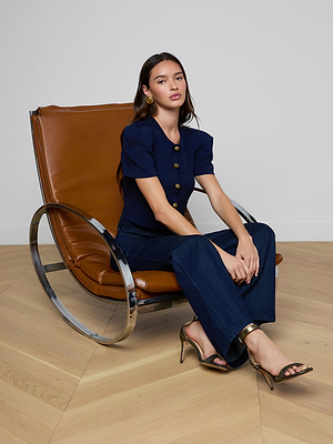 A woman with long dark hair sits on a modern brown leather chair, wearing the Britt Statement Shoulder Cardigan, dark jeans, heeled sandals, and gold earrings—creating a striking look against light wooden floors and a white wall.