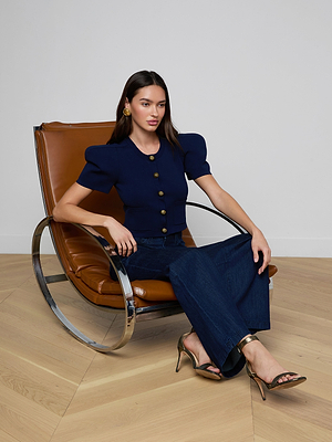 A woman sits on a modern chair, wearing the Britt Statement Shoulder Cardigan with puffed sleeves and gold buttons, wide-leg dark jeans, heeled sandals, and gold hoop earrings. Her long brown hair completes this standout look.