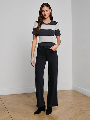 A woman with long brown hair and a neutral expression stands indoors on a wooden floor, wearing the Rosemond Bodysuit with short sleeves and wide stripes, paired with black high-waisted wide-leg jeans and black boots.