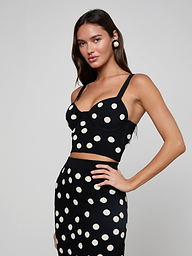 A woman with long brown hair wears the Tula Knit Bra Top, a black compression knit crop top with large jacquard polka dots, paired with a matching high-waisted skirt. She poses against a plain light gray background, hand on hip.