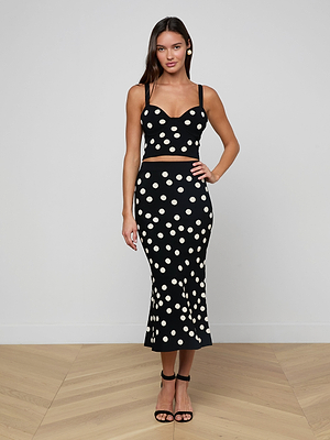 A woman stands indoors against a white wall, wearing the Tula Knit Bra Top with a high-waisted, mid-length black polka dot skirt and black heels. Her straight brown hair complements the chic two-piece outfit.
