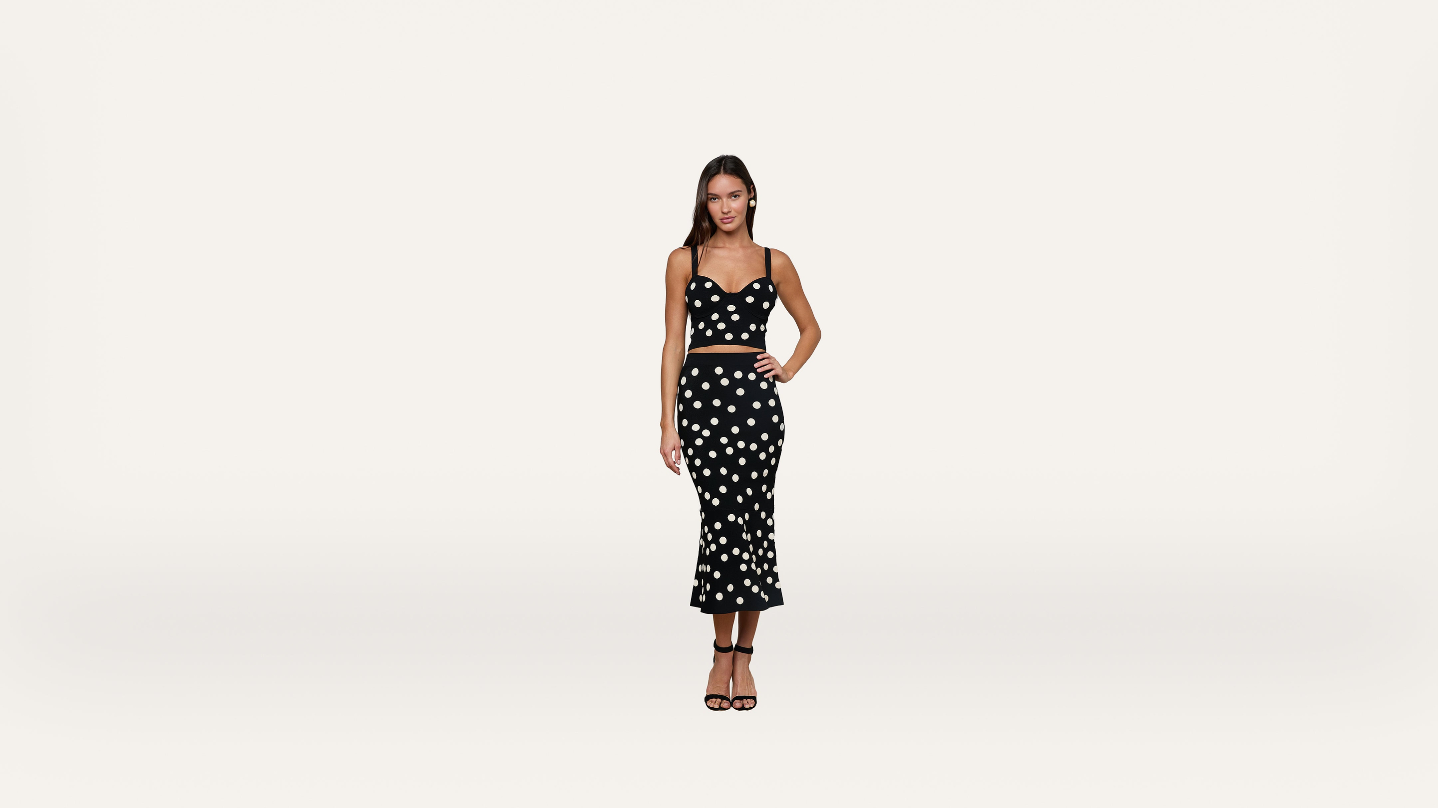 A woman stands against a plain white background, wearing a black crop top and matching midi skirt with white polka dots, and black high-heeled sandals.