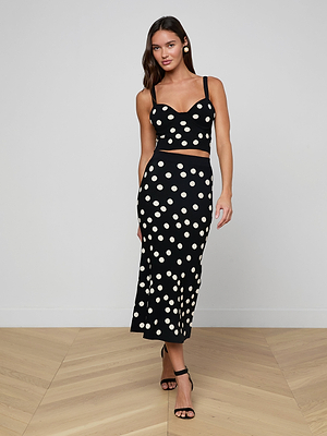 A woman with long dark hair poses indoors against a plain light wall and wooden floor, wearing the Tula Knit Bra Top—a black, corset-style crop top—paired with a matching high-waisted jacquard polka dot skirt and black strappy heels.