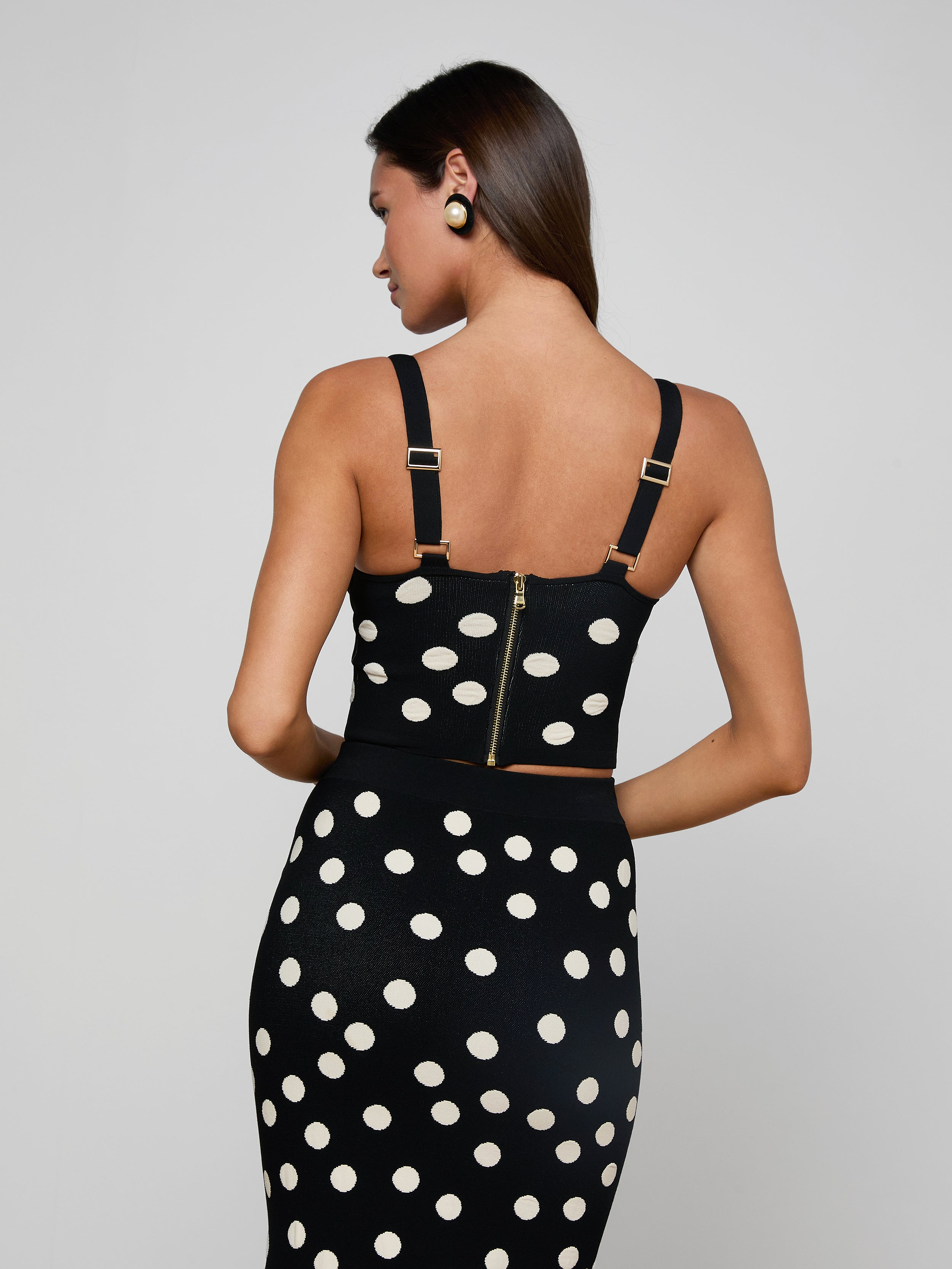 A woman stands with her back to the camera, wearing the Tula Knit Bra Top in black with jacquard polka dots and gold zipper details, paired with a matching skirt. Her straight dark hair and black-and-gold earrings complete her chic look.