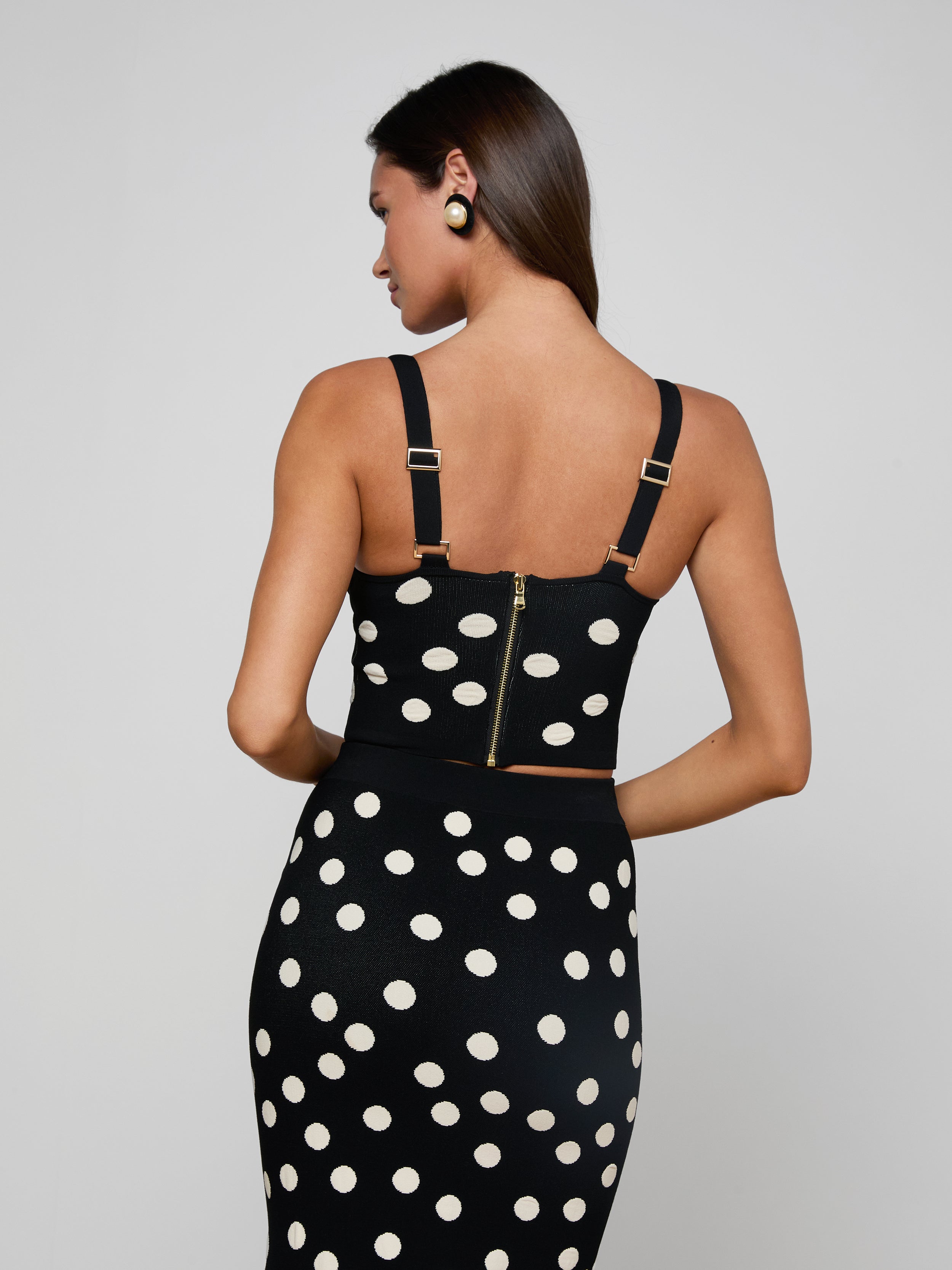 A woman stands with her back to the camera, wearing the Tula Knit Bra Top in black with jacquard polka dots and gold zipper details, paired with a matching skirt. Her straight dark hair and black-and-gold earrings complete her chic look.
