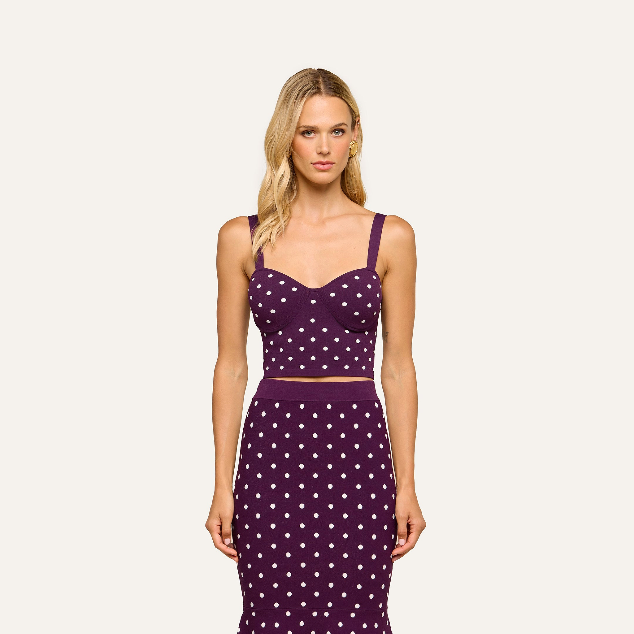A woman with long blonde hair wears the Tula Knit Bra Top—a sleeveless, dark purple crop top featuring jacquard polka dots—paired with a matching skirt, standing against a plain light background.