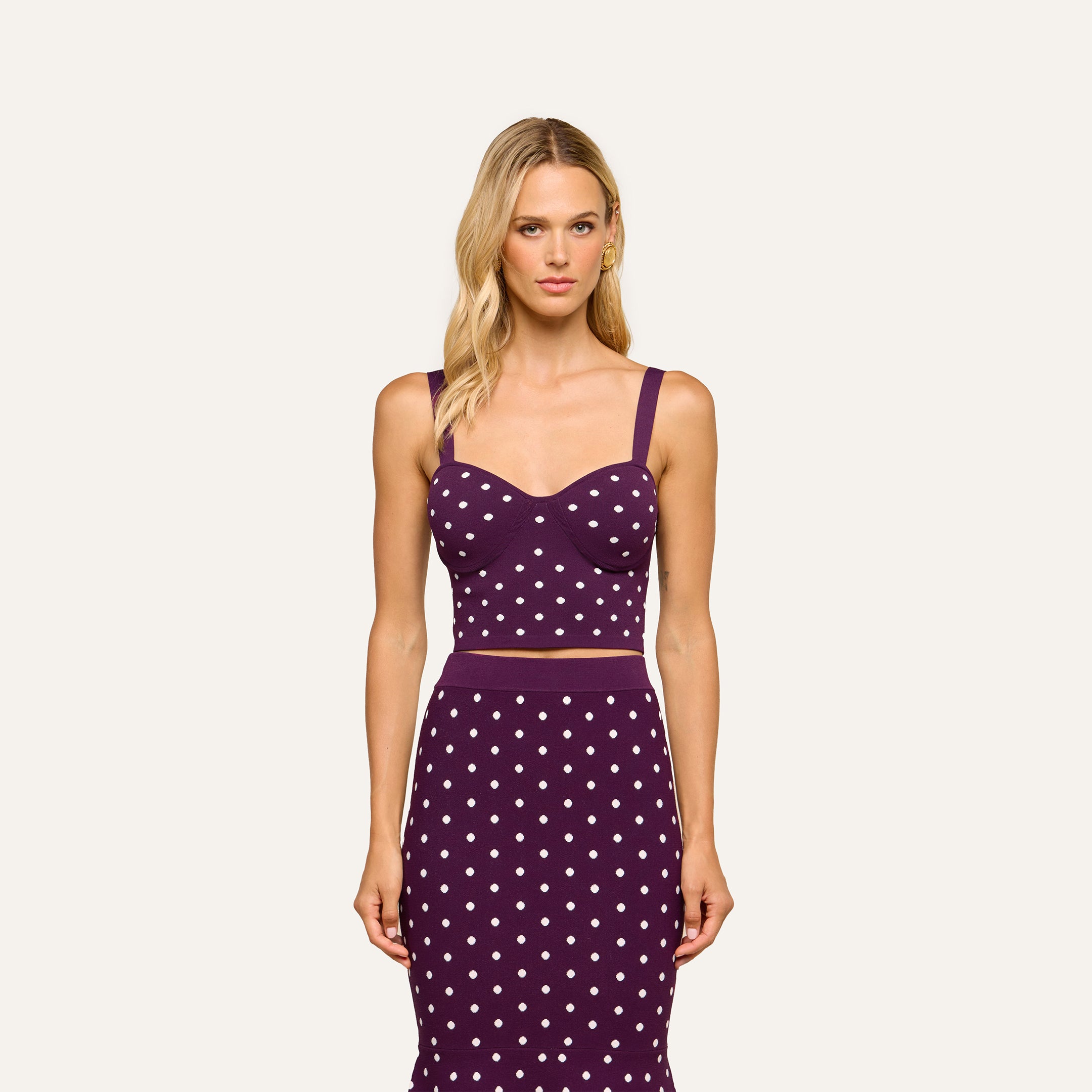 A woman with long blonde hair wears the Tula Knit Bra Top—a sleeveless, dark purple crop top featuring jacquard polka dots—paired with a matching skirt, standing against a plain light background.