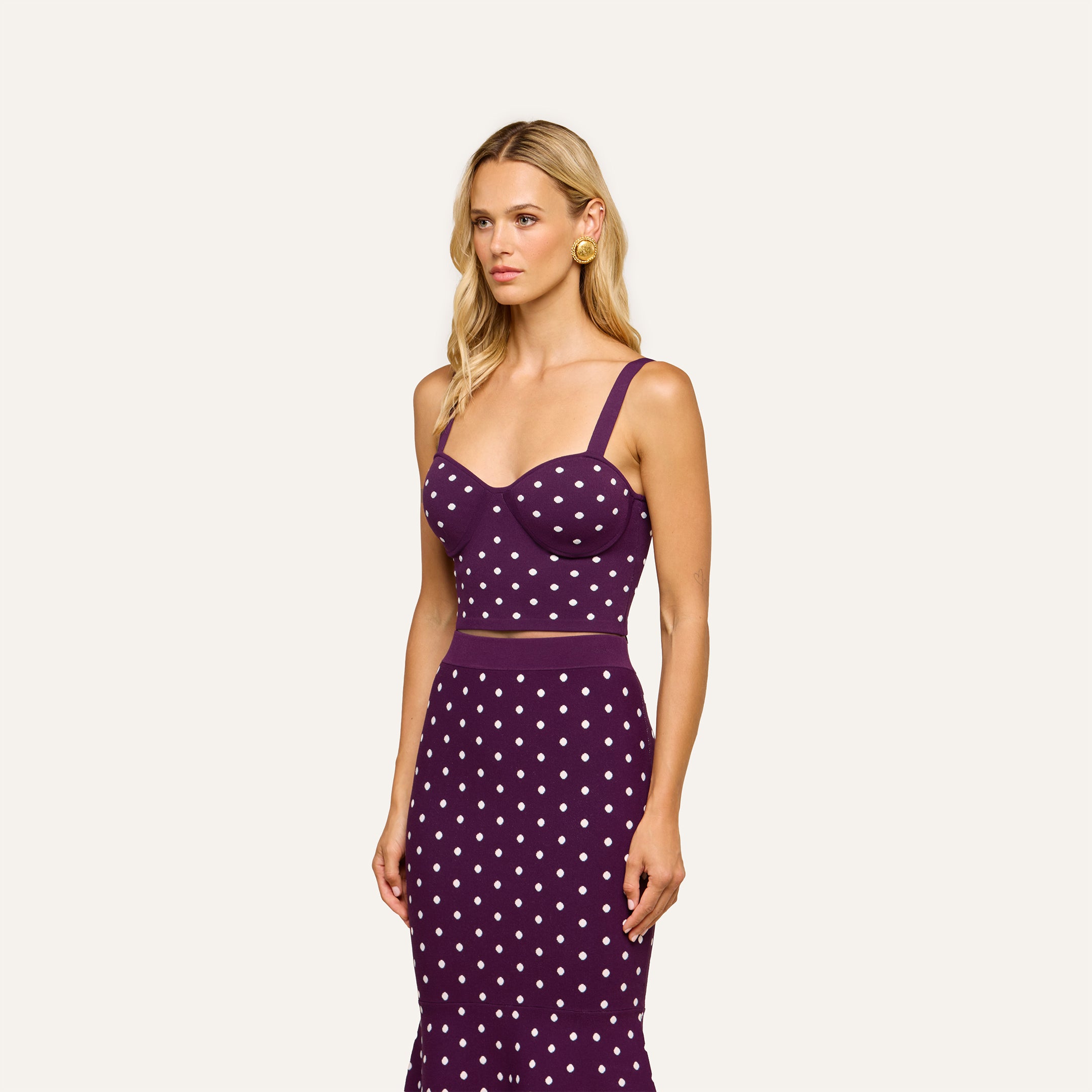 A woman with long blonde hair wears the Tula Knit Bra Top—a corset-style purple crop top—paired with a matching jacquard polka dot skirt, standing against a plain light background.