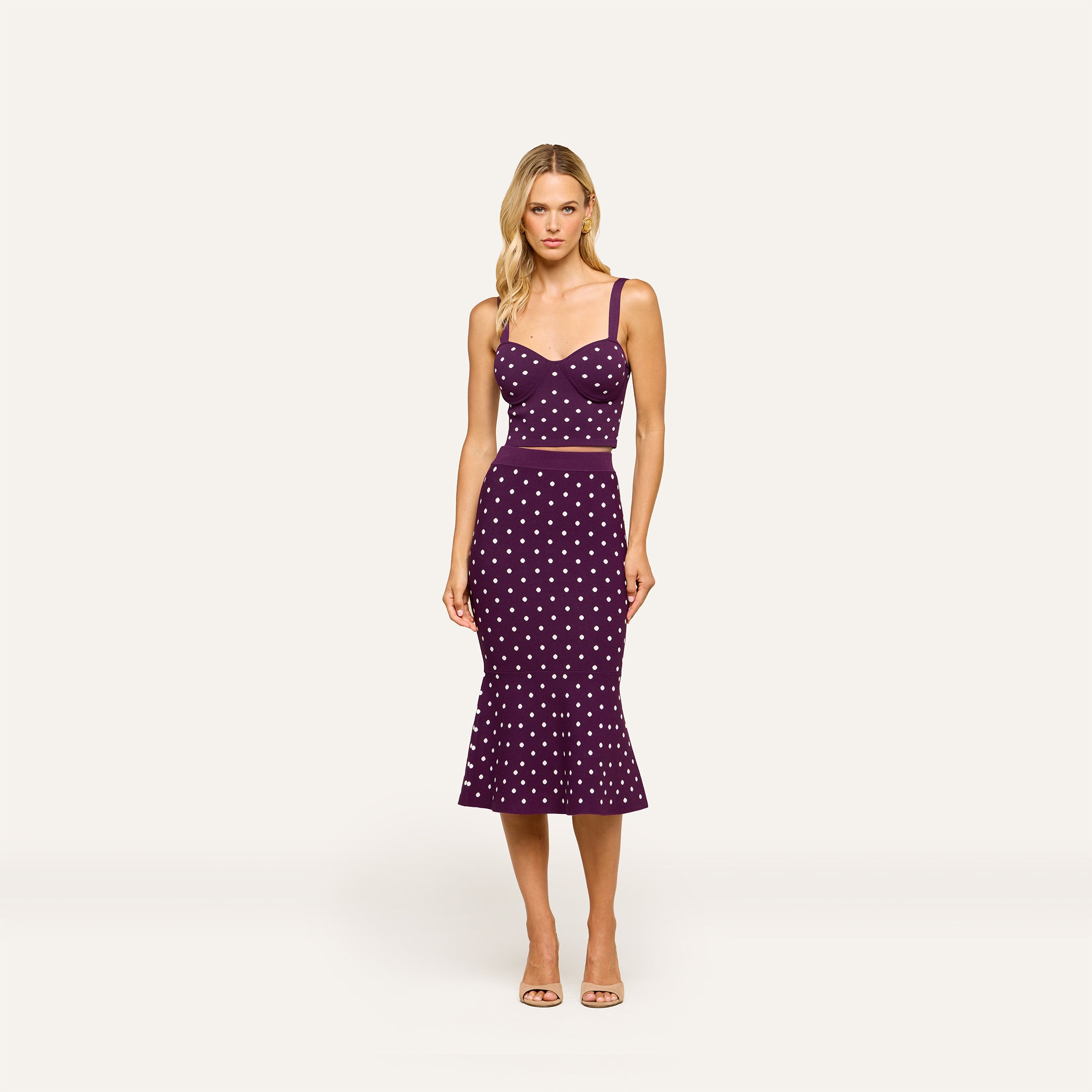 A woman stands against a plain light background wearing the Tula Knit Bra Top, a purple sleeveless crop top with corset-style details and jacquard polka dot pattern, paired with a matching high-waisted, mid-length skirt.