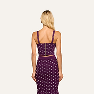 A woman with long blonde hair stands facing away, wearing the Tula Knit Bra Top—a purple crop top with jacquard polka dots, thick straps, and a visible gold zipper on the corset-style bodice—paired with a matching skirt.