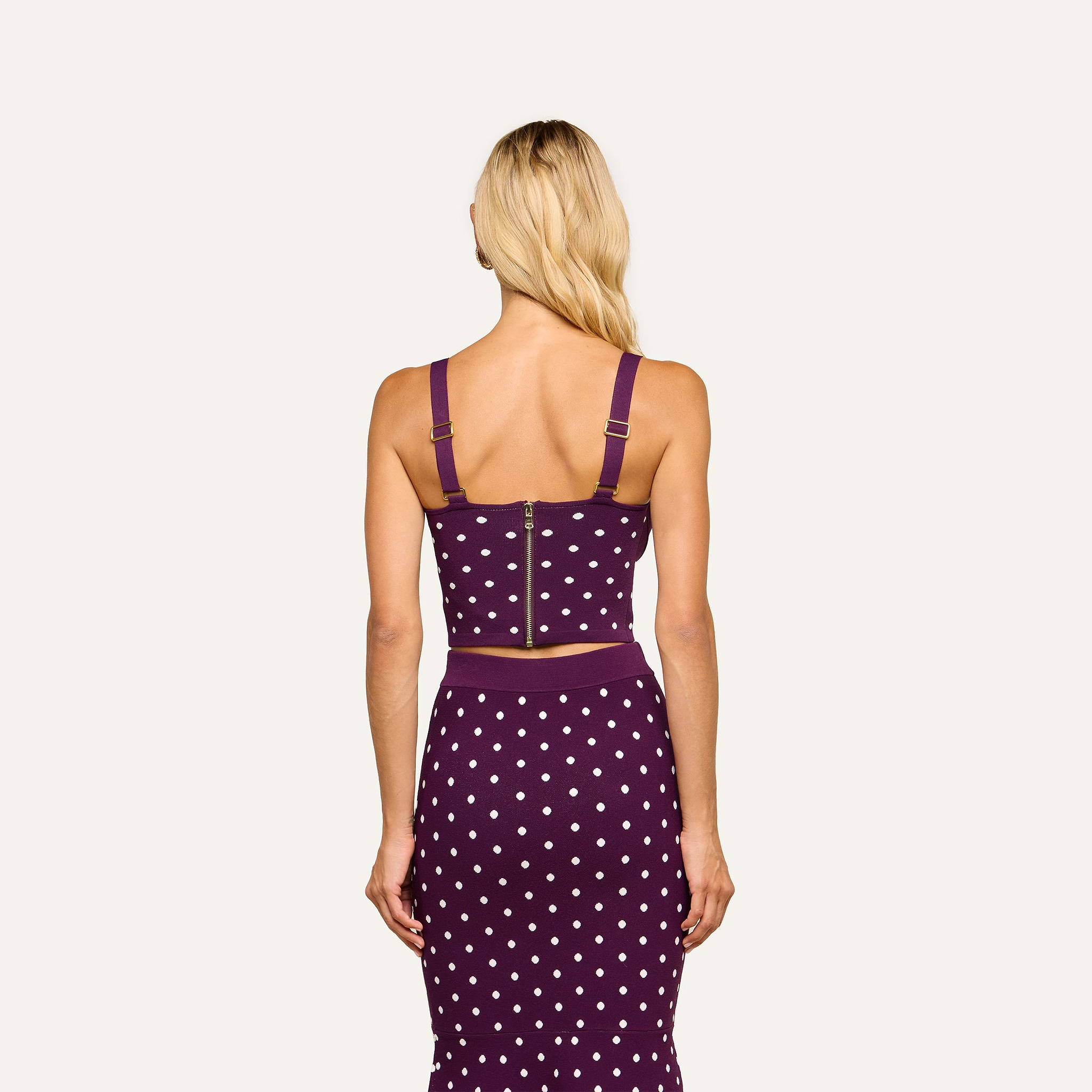 A woman with long blonde hair stands facing away, wearing the Tula Knit Bra Top—a purple crop top with jacquard polka dots, thick straps, and a visible gold zipper on the corset-style bodice—paired with a matching skirt.