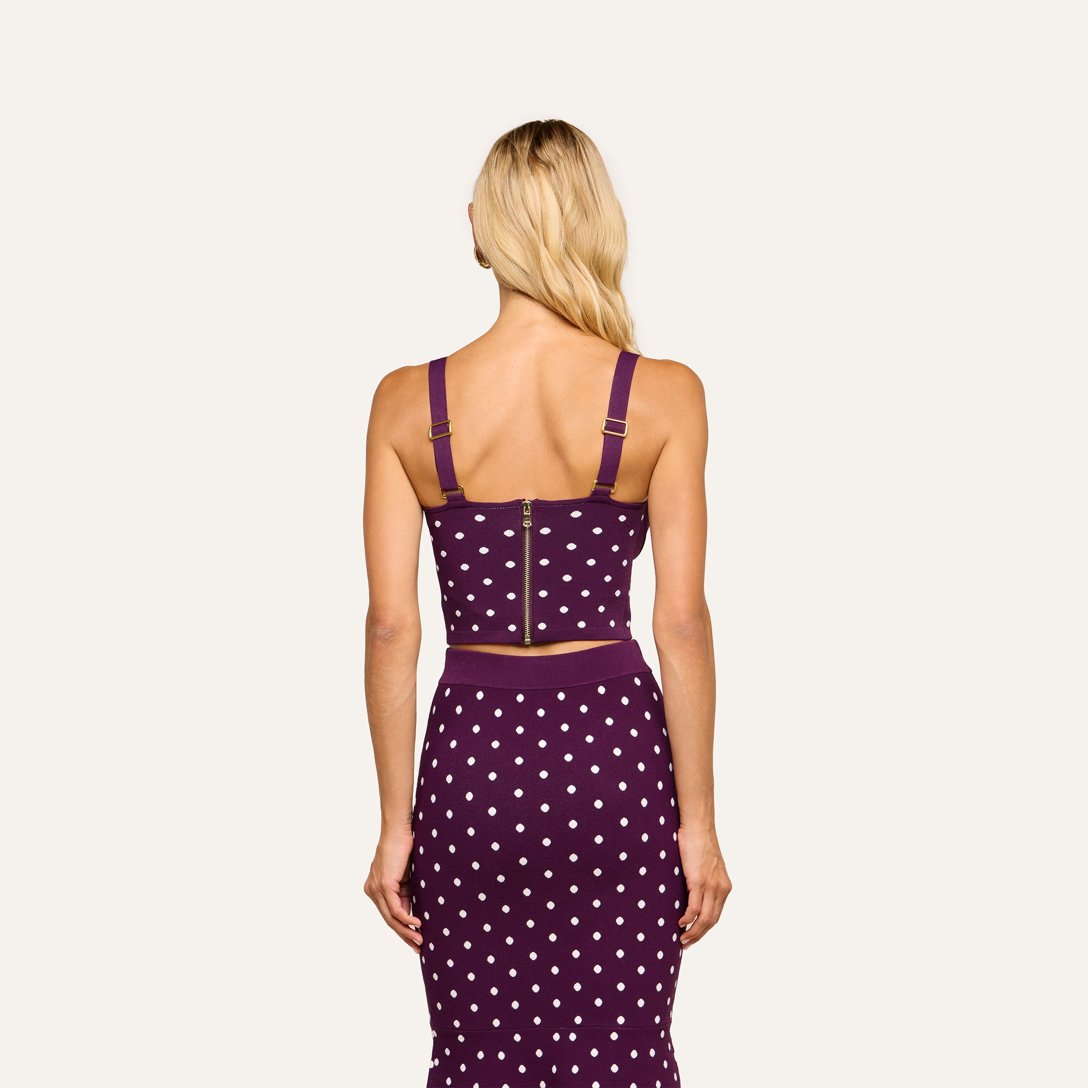 A woman with long blonde hair stands facing away, wearing the Tula Knit Bra Top—a purple crop top with jacquard polka dots, thick straps, and a visible gold zipper on the corset-style bodice—paired with a matching skirt.