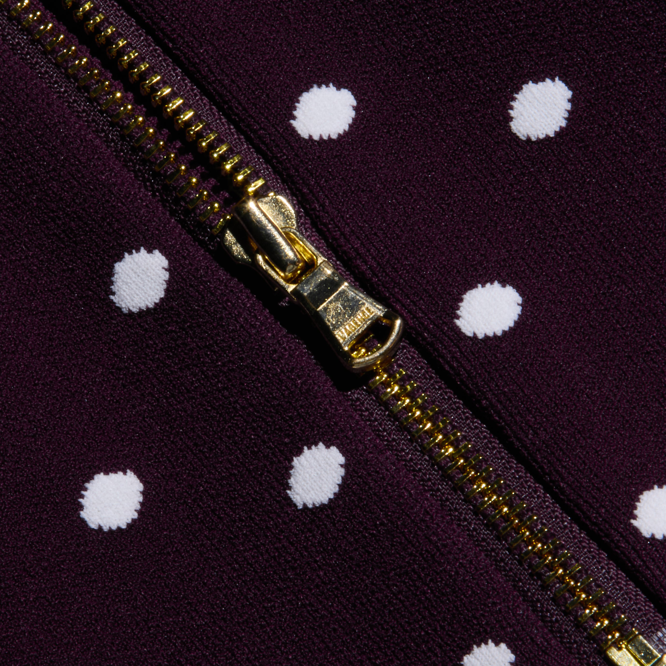 A close-up of the Tula Knit Bra Top’s gold zipper on dark purple fabric featuring evenly spaced white jacquard polka dots.